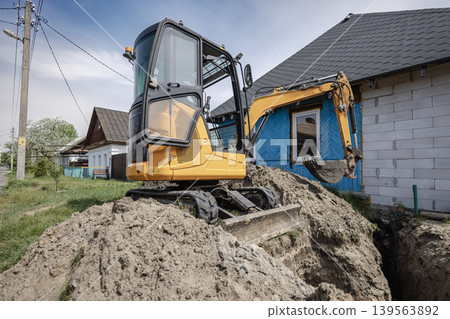 A yellow excavator digs a trench beside a house in a suburban area. The machine sits on a pile of dirt with clear skies above and other houses in the background A yellow excavator digs a trench beside a house in a suburban area. The machine sits on a pile of dirt with clear skies above and other houses in the background 139563892