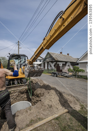 A worker operates an excavator to dig a trench beside houses. The machine lifts dirt in a residential area on a clear day with power lines overhead A worker operates an excavator to dig a trench beside houses. The machine lifts dirt in a residential area on a clear day with power lines overhead 139563895