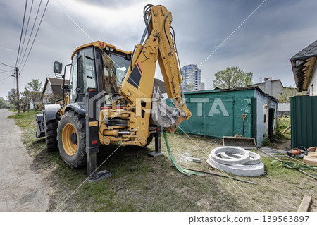 A backhoe loader is parked by the road in a residential area. It has a shovel attached and is next to a green shed with nearby buildings in the background 139563897