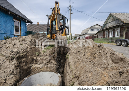 Workers operate a digger to create a trench in a rural setting. Nearby houses and utility poles are visible in the background under an overcast sky Workers operate a digger to create a trench in a rural setting. Nearby houses and utility poles are visible in the background under an overcast sky 139563898