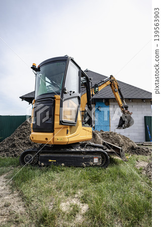 A small excavator is digging soil at a construction site. The machine is next to a building that is being developed. There are dirt piles around the area 139563903