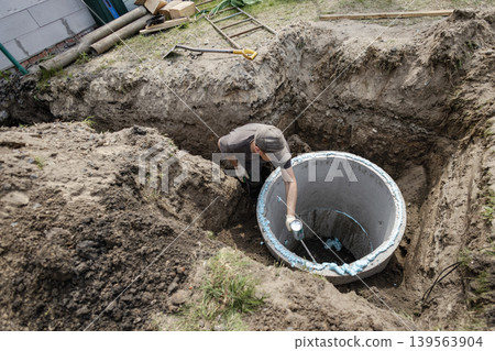 A person works at a construction site in a backyard. They are filling a concrete pit with materials. Dirt and tools are scattered around A person works at a construction site in a backyard. They are filling a concrete pit with materials. Dirt and tools are scattered around 139563904