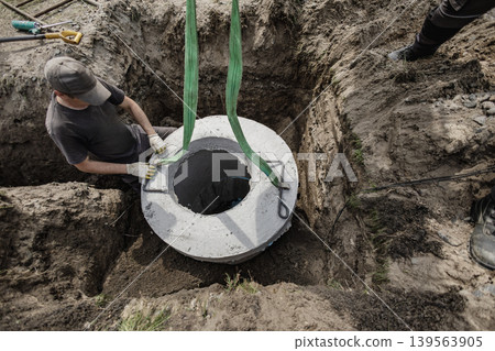 A worker is placing a concrete well cover into a dug pit at a construction site. Tools and soil are around him as he focuses on the task A worker is placing a concrete well cover into a dug pit at a construction site. Tools and soil are around him as he focuses on the task 139563905