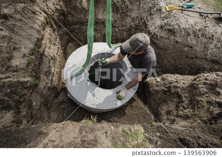 A worker places a concrete septic tank into a hole at a construction site. The worker uses straps for lifting while surrounded by dirt and tools A worker places a concrete septic tank into a hole at a construction site. The worker uses straps for lifting while surrounded by dirt and tools 139563906