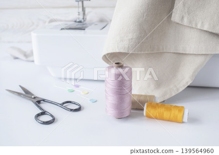 Sewing supplies on a white table with a sewing machine in the background. Sewing machine with fabric and sewing accessories on a white table. Sewing supplies on a white table with a sewing machine in the background. Sewing machine with fabric and sewing accessories on a white table. 139564960