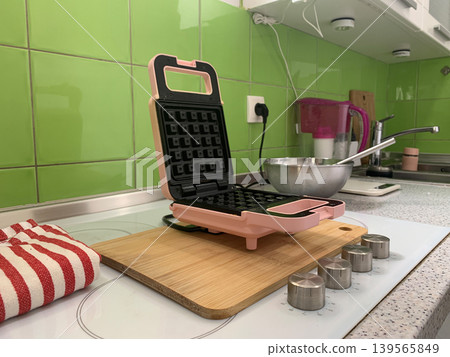 Angled left view of an open pink waffle maker on a wooden board with a striped towel nearby. Angled left view of an open pink waffle maker on a wooden board with a striped towel nearby. 139565849
