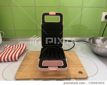 High angle centered view of an open pink waffle maker on a wooden board with a striped towel. High angle centered view of an open pink waffle maker on a wooden board with a striped towel. 139565850
