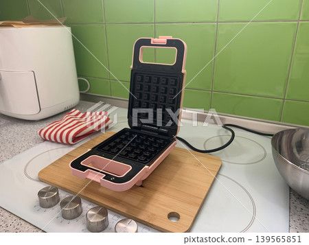 High angle right view of an open pink waffle maker and a white appliance on a kitchen counter. High angle right view of an open pink waffle maker and a white appliance on a kitchen counter. 139565851