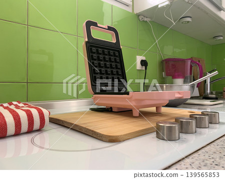 Low angle left view of a pink mini waffle maker plugged into a kitchen wall. Low angle left view of a pink mini waffle maker plugged into a kitchen wall. 139565853