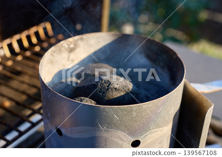 Charcoal briquettes smoke inside a metal chimney starter on an outdoor grill Charcoal briquettes smoke inside a metal chimney starter on an outdoor grill 139567108