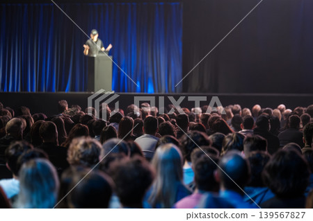 Audience watching speaker on stage at conference presentation with blue curtain and auditorium crowd Audience watching speaker on stage at conference presentation with blue curtain and auditorium crowd 139567827