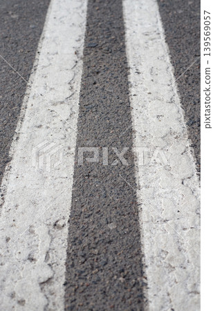 Double stripes of white road markings with cracks on dark asphalt. Gray road surface texture and details of a city street with signs of wear and tear. Double stripes of white road markings with cracks on dark asphalt. Gray road surface texture and details of a city street with signs of wear and tear. 139569057