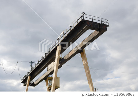 Industrial gantry crane structure against a dramatic cloudy sky, viewed from below with weathered metal beams and operator cabin 139569062