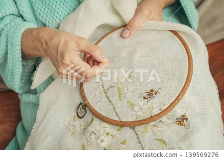 Hands stitch floral embroidery on fabric held in hoop, flower pattern, wrinkled hands close-up. Concept of pleasant home leisure, embroidery craft for hobby, handmade product marketing Hands stitch floral embroidery on fabric held in hoop, flower pattern, wrinkled hands close-up. Concept of pleasant home leisure, embroidery craft for hobby, handmade product marketing 139569276