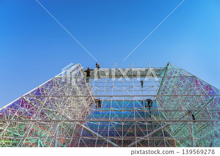 Workers assemble metal structure on geometric glass facade under blue sky with wide. Concept of structure, architecture, construction, engineering, teamwork, urban development. Copy space Workers assemble metal structure on geometric glass facade under blue sky with wide. Concept of structure, architecture, construction, engineering, teamwork, urban development. Copy space 139569278