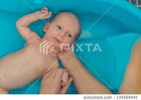 Adorable newborn baby bathing in a blue tub at home with gentle care. Cute infant hygiene, parenting, wellness and baby care routine concept with tenderness, comfort and family love Adorable newborn baby bathing in a blue tub at home with gentle care. Cute infant hygiene, parenting, wellness and baby care routine concept with tenderness, comfort and family love 139569445