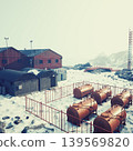 Nestled in a snowy terrain, a collection of red and gray buildings stands near large fuel tanks. The scene is serene, with rocky mountains in the backdrop under a pale sky. 139569820