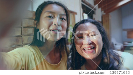 Taking mother and daughter leaning close wearing yellow top and light top at home, holding phone Taking mother and daughter leaning close wearing yellow top and light top at home, holding phone 139571653