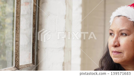 Gazing mature woman wearing Santa hat by metal window frame at home, brick wall, copy space Gazing mature woman wearing Santa hat by metal window frame at home, brick wall, copy space 139571683
