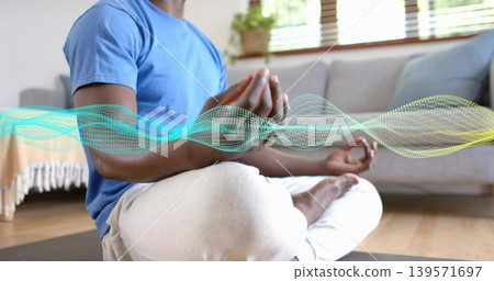 Meditating man wearing blue t-shirt, gray sweats crosslegged on yoga mat at home, with digital wave Meditating man wearing blue t-shirt, gray sweats crosslegged on yoga mat at home, with digital wave 139571697