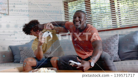 Playing, two men wearing orange on gray sofa at home, holding white controllers, sharing popcorn Playing, two men wearing orange on gray sofa at home, holding white controllers, sharing popcorn 139571717