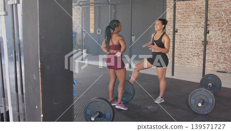 Talking two women wearing maroon and black gear exchanging cues in lifting gym with 20KG plates Talking two women wearing maroon and black gear exchanging cues in lifting gym with 20KG plates 139571727