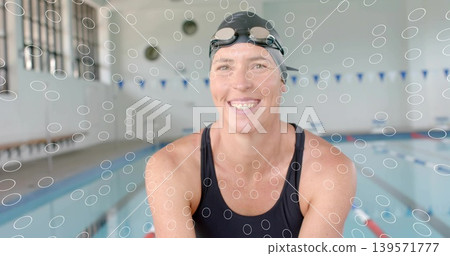 Smiling woman swimmer leaning on pool edge, wearing black one-piece swimsuit, swim cap, lane ropes Smiling woman swimmer leaning on pool edge, wearing black one-piece swimsuit, swim cap, lane ropes 139571777