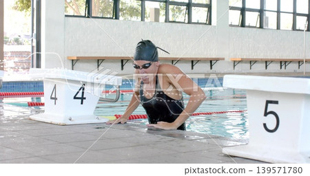 Emerging adult swimmer pushing onto pool deck at blocks-4-and-5, wearing black cap goggles onepiece Emerging adult swimmer pushing onto pool deck at blocks-4-and-5, wearing black cap goggles onepiece 139571780