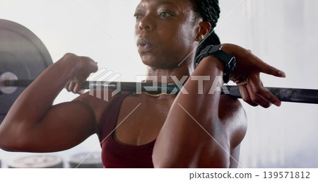 Holding loaded barbell across front shoulders, woman training in gym wearing maroon top, smartwatch Holding loaded barbell across front shoulders, woman training in gym wearing maroon top, smartwatch 139571812