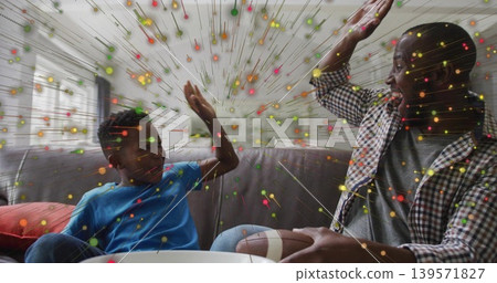 High-fiving dad and son on gray sofa at home, checked-shirt-blue-tee, football-and-bowl, confetti 139571827