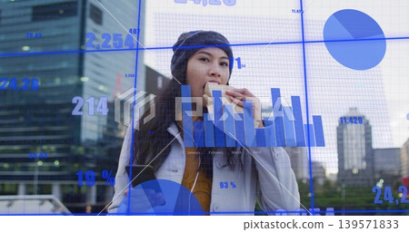 Biting sandwich, woman in knit beanie and coat checking smartphone in plaza, with finance charts 139571833