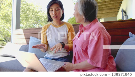 Showing two Asian women gesturing over laptop on porch, wearing raglan tee, pink shirt, smartwatch Showing two Asian women gesturing over laptop on porch, wearing raglan tee, pink shirt, smartwatch 139571940