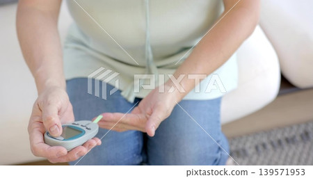 Holding Asian woman wearing light green top, blue jeans checking glucose on sofa, with meter Holding Asian woman wearing light green top, blue jeans checking glucose on sofa, with meter 139571953