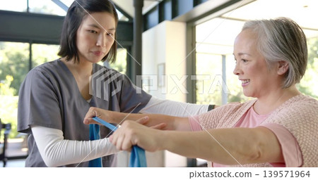 Performing caregiver and senior patient pulling blue resistance band in rehab room, scrubs, windows 139571964