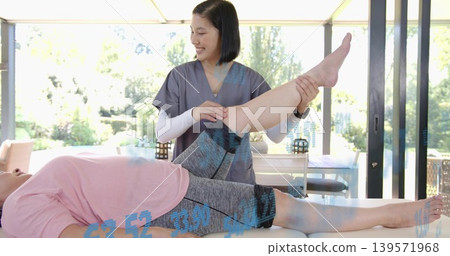 Stabilizing therapist in gray scrubs lifting patient's bent leg on treatment table, pink tee Stabilizing therapist in gray scrubs lifting patient's bent leg on treatment table, pink tee 139571968