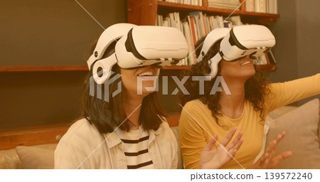 Reaching, smiling women wearing white VR headsets on sofa, striped and yellow tops near bookshelf Reaching, smiling women wearing white VR headsets on sofa, striped and yellow tops near bookshelf 139572240