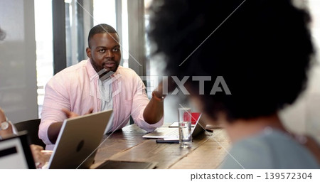 Gesturing man in pink shirt over gray tee leading meeting at conference table, with laptops papers 139572304
