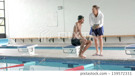 Coaching coach holding tablet leaning toward swimmer on block 2 at pool, lanes, cap, copy space Coaching coach holding tablet leaning toward swimmer on block 2 at pool, lanes, cap, copy space 139572331
