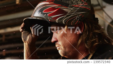 Holding mature welder checking helmet with red graphics in workshop, in dark shirt, sparks Holding mature welder checking helmet with red graphics in workshop, in dark shirt, sparks 139572340
