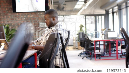 Typing man wearing plaid shirt focusing on laptop at open-plan office, wall monitor visible Typing man wearing plaid shirt focusing on laptop at open-plan office, wall monitor visible 139572343