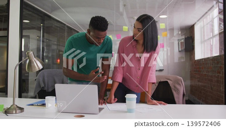 Leaning woman in pink blouse and man in green t-shirt reviewing laptop and documents at office Leaning woman in pink blouse and man in green t-shirt reviewing laptop and documents at office 139572406