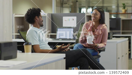 Chatting colleagues at office desks, woman in pink blouse holding takeout using chopsticks, laptop Chatting colleagues at office desks, woman in pink blouse holding takeout using chopsticks, laptop 139572409