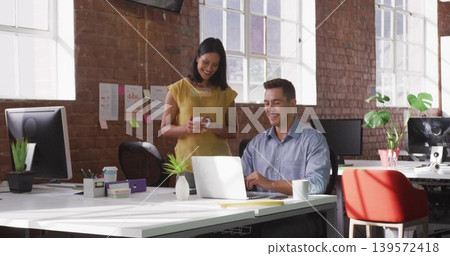 Standing woman in mustard blouse holding white mug, smiling at man using silver laptop at office Standing woman in mustard blouse holding white mug, smiling at man using silver laptop at office 139572418