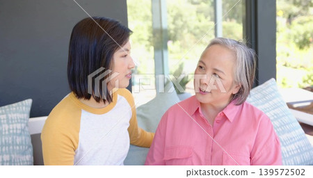 Leaning yellow-sleeved daughter placing arm behind mother on sofa, pillows, garden beyond glass 139572502
