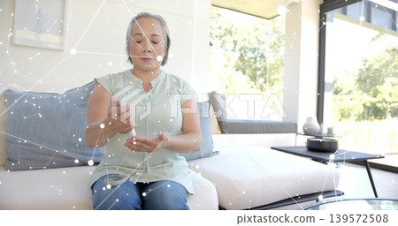 Dispensing senior Asian woman mint blouse, jeans pressing pump bottle on sofa at home, with trees Dispensing senior Asian woman mint blouse, jeans pressing pump bottle on sofa at home, with trees 139572508
