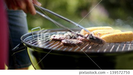Grilling burger patties and corn on charcoal kettle grill in backyard, wearing denim and red top Grilling burger patties and corn on charcoal kettle grill in backyard, wearing denim and red top 139572750