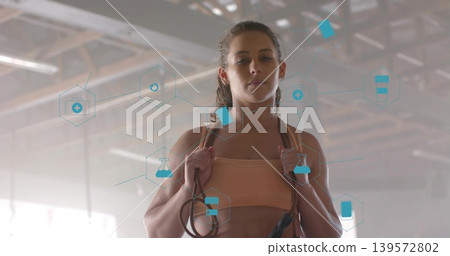 Standing woman in peach sports bra holding jump rope at gym, with blue AR health icons Standing woman in peach sports bra holding jump rope at gym, with blue AR health icons 139572802