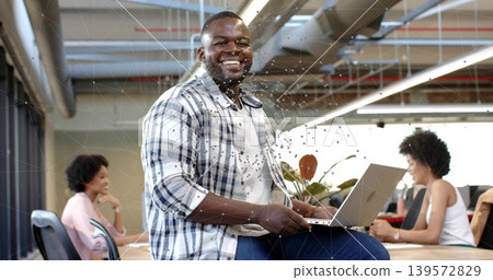 Smiling man wearing plaid shirt sitting on long wooden table holding laptop, working in open office Smiling man wearing plaid shirt sitting on long wooden table holding laptop, working in open office 139572829