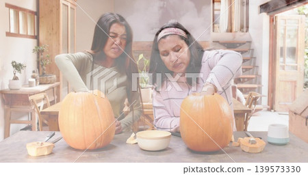Carving two women leaning over table in cozy kitchen wearing headband and using pumpkins and tools Carving two women leaning over table in cozy kitchen wearing headband and using pumpkins and tools 139573330