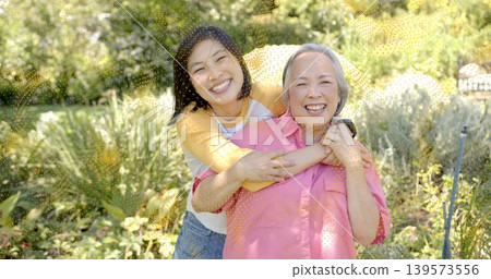 Hugging daughter holding mother in sunny garden, wearing casual clothes with smartwatch necklace Hugging daughter holding mother in sunny garden, wearing casual clothes with smartwatch necklace 139573556